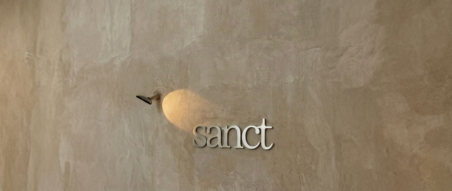 The Sanct wordmark mounted on a warm plaster wall, lit by a soft pool of light